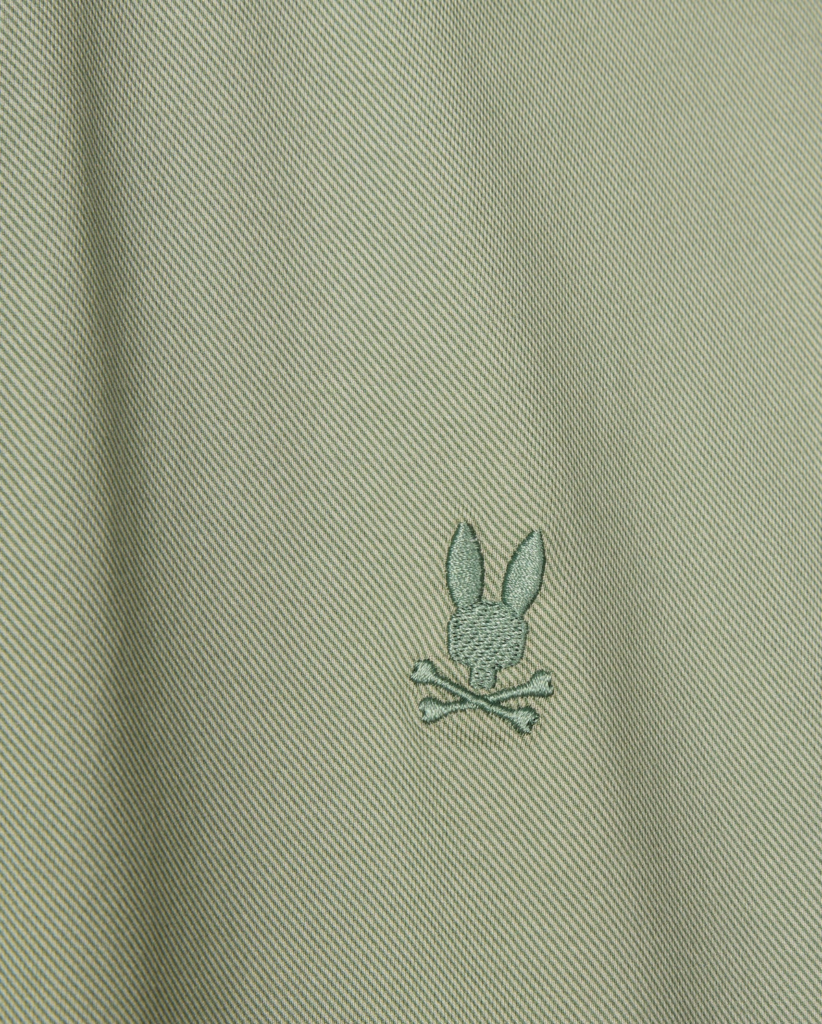 Close-up of the Psycho Bunny's Men's Brendan Jersey Shirt in light green Pima cotton, showcasing a subtle diagonal texture. The shirt features a small embroidered chest Bunny logo, depicting a rabbit head above two crossed bones, centered on the fabric.