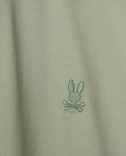 Close-up of the Psycho Bunny's Men's Brendan Jersey Shirt in light green Pima cotton, showcasing a subtle diagonal texture. The shirt features a small embroidered chest Bunny logo, depicting a rabbit head above two crossed bones, centered on the fabric.