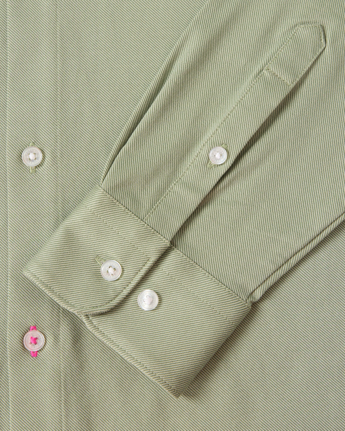 Zoom in on the sleeve of the MENS BRENDAN JERSEY SHIRT - B6C749D200 by Psycho Bunny, featuring a green, herringbone-patterned design. The sleeve showcases a cuff with three white buttons, crafted from Pima cotton jersey fabric that offers a subtle texture. A unique detail is highlighted by one buttonhole stitched with pink thread. Sophisticated contrast piping along the edges enhances its elegant appeal.
