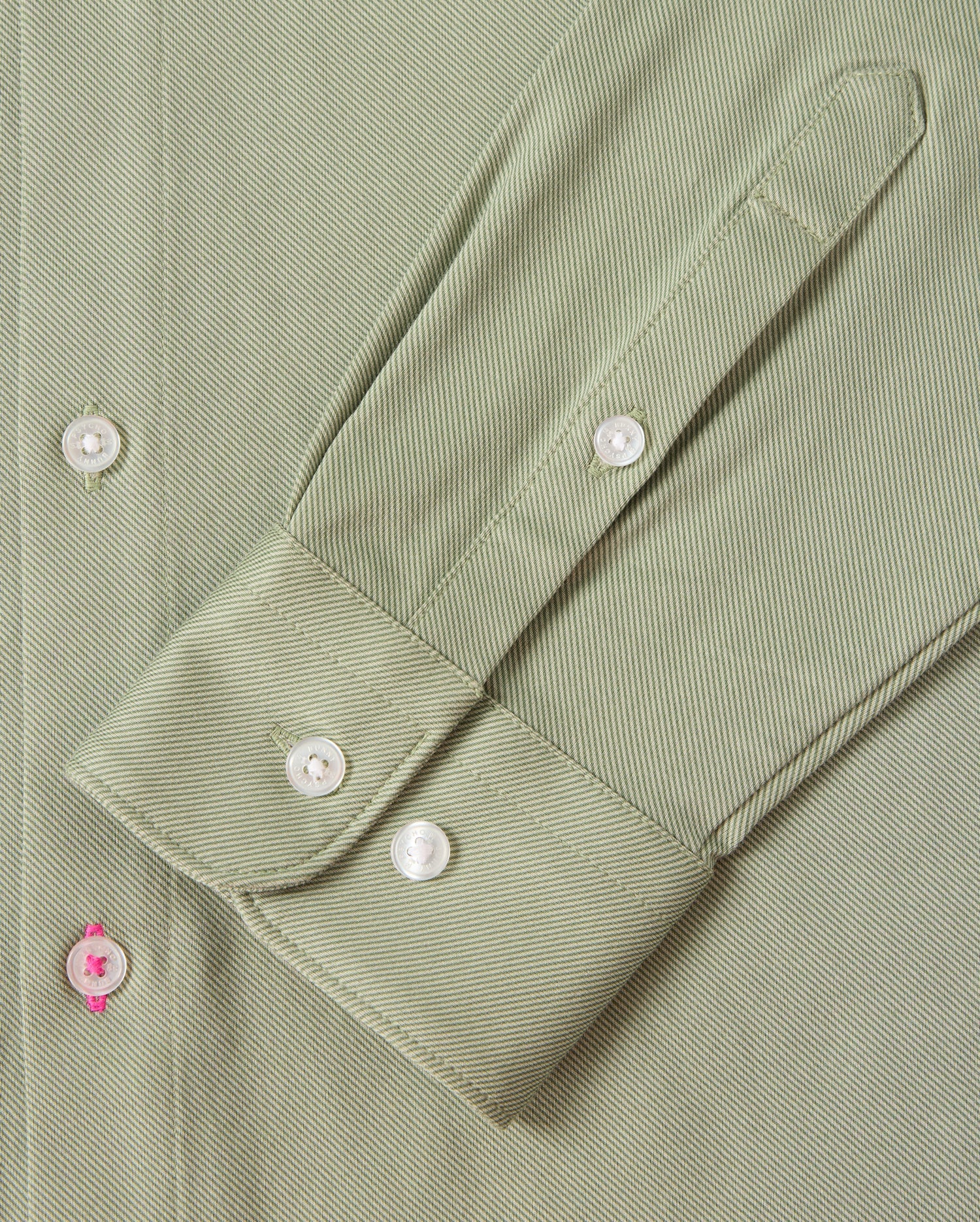 Zoom in on the sleeve of the MENS BRENDAN JERSEY SHIRT - B6C749D200 by Psycho Bunny, featuring a green, herringbone-patterned design. The sleeve showcases a cuff with three white buttons, crafted from Pima cotton jersey fabric that offers a subtle texture. A unique detail is highlighted by one buttonhole stitched with pink thread. Sophisticated contrast piping along the edges enhances its elegant appeal.