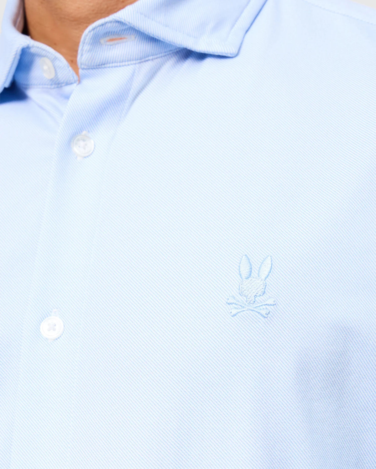 Close-up of a person wearing the light blue Psycho Bunny MENS BRENDAN JERSEY SHIRT - B6C749D200, featuring a subtle Bunny and crossbones logo embroidered on the chest. This shirt, crafted from textured Pima cotton, exudes a relaxed vibe with its top button left stylishly unfastened.