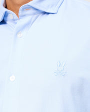 Close-up of a person wearing the light blue Psycho Bunny MENS BRENDAN JERSEY SHIRT - B6C749D200, featuring a subtle Bunny and crossbones logo embroidered on the chest. This shirt, crafted from textured Pima cotton, exudes a relaxed vibe with its top button left stylishly unfastened.