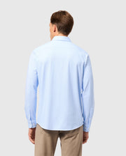 A man with short brown hair is wearing the MENS BRENDAN JERSEY SHIRT - B6C749D200, a light blue button-up crafted from luxurious Pima cotton by Psycho Bunny, along with beige pants. He stands with his back to the camera against a plain, light gray background.