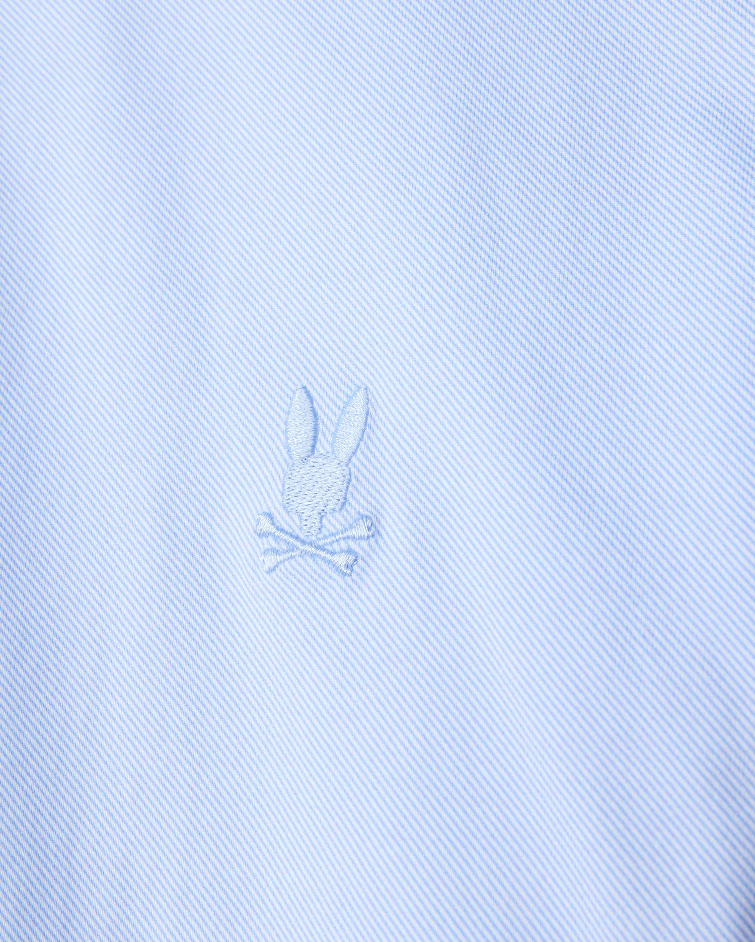 Close-up of the light blue stretch jersey fabric of the Psycho Bunny MENS BRENDAN JERSEY SHIRT - B6C749D200, highlighting the embroidered chest Bunny logo featuring a rabbit head with crossed bones beneath it. The design is subtle, with the logo in a slightly darker shade of blue than the Pima cotton fabric.