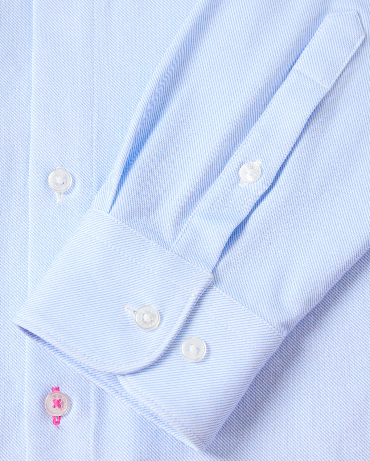 Close-up of the sleeve on the Psycho Bunny MENS BRENDAN JERSEY SHIRT - B6C749D200, crafted in a soft light blue pinstripe from premium Pima cotton. It features a neatly buttoned cuff with a striking pink buttonhole accent against smooth white buttons. The shirt is laid flat, showcasing the sleeve's precise stitching and button detailing.