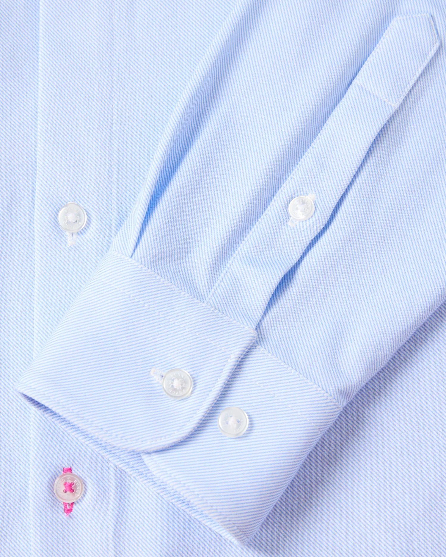 Close-up of the sleeve on the Psycho Bunny MENS BRENDAN JERSEY SHIRT - B6C749D200, crafted in a soft light blue pinstripe from premium Pima cotton. It features a neatly buttoned cuff with a striking pink buttonhole accent against smooth white buttons. The shirt is laid flat, showcasing the sleeve's precise stitching and button detailing.