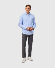 A man stands against a light gray background, wearing a cozy Psycho Bunny MENS BROOK STRIPED JERSEY SHIRT - B6C750D200 in light blue Pima cotton, paired with dark gray pants and white sneakers. He gazes forward with his hands clasped in front of him.