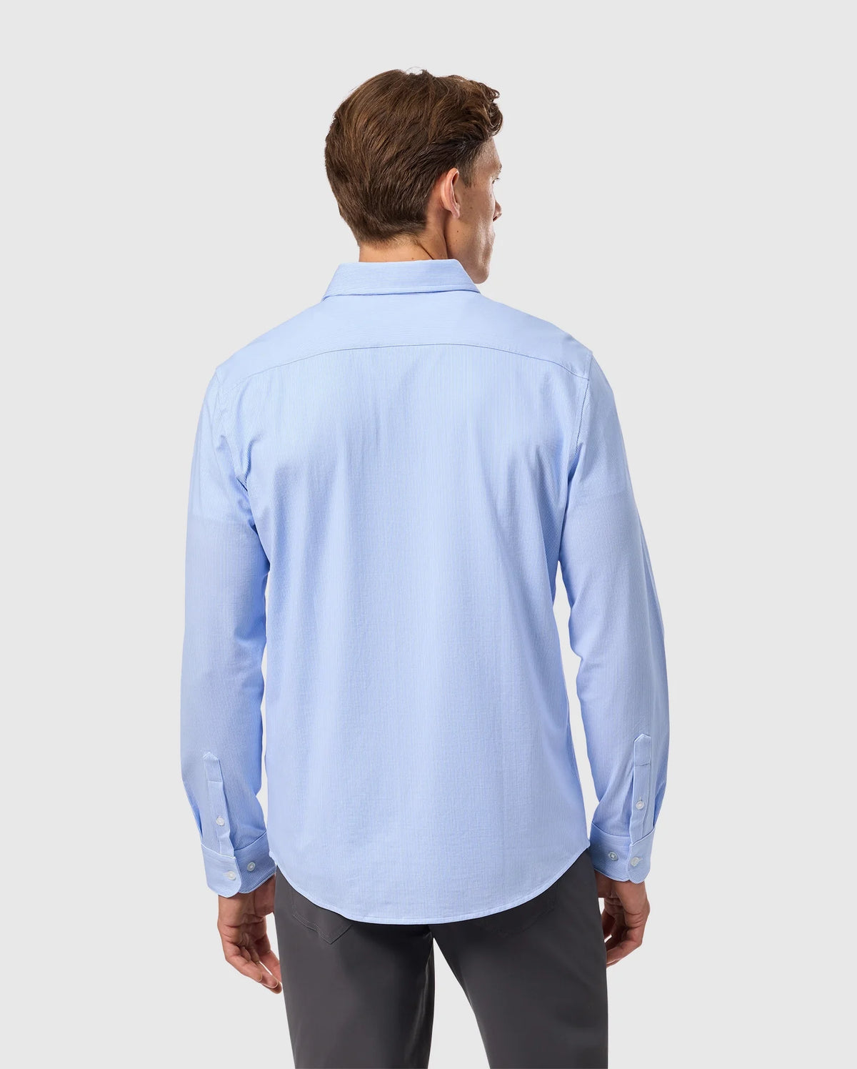 A man wearing a Psycho Bunny light blue striped jersey shirt made of Pima cotton, identified as product B6C750D200, with long sleeves and gray pants, stands facing away from the camera against a white background.