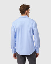 A man wearing a Psycho Bunny light blue striped jersey shirt made of Pima cotton, identified as product B6C750D200, with long sleeves and gray pants, stands facing away from the camera against a white background.