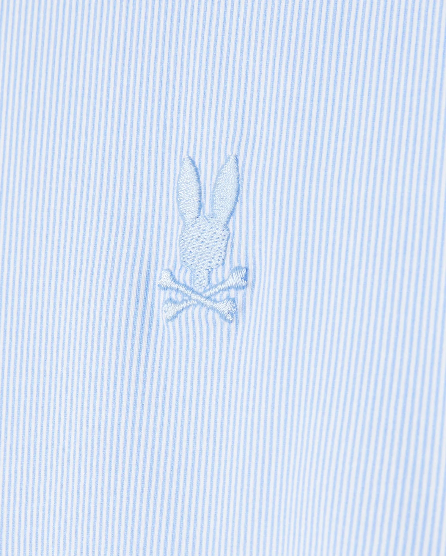 Close-up of the Psycho Bunny men's Brook striped jersey shirt in light blue and white, made from soft Pima cotton and featuring an embroidered emblem of a bunny head above crossed bones.