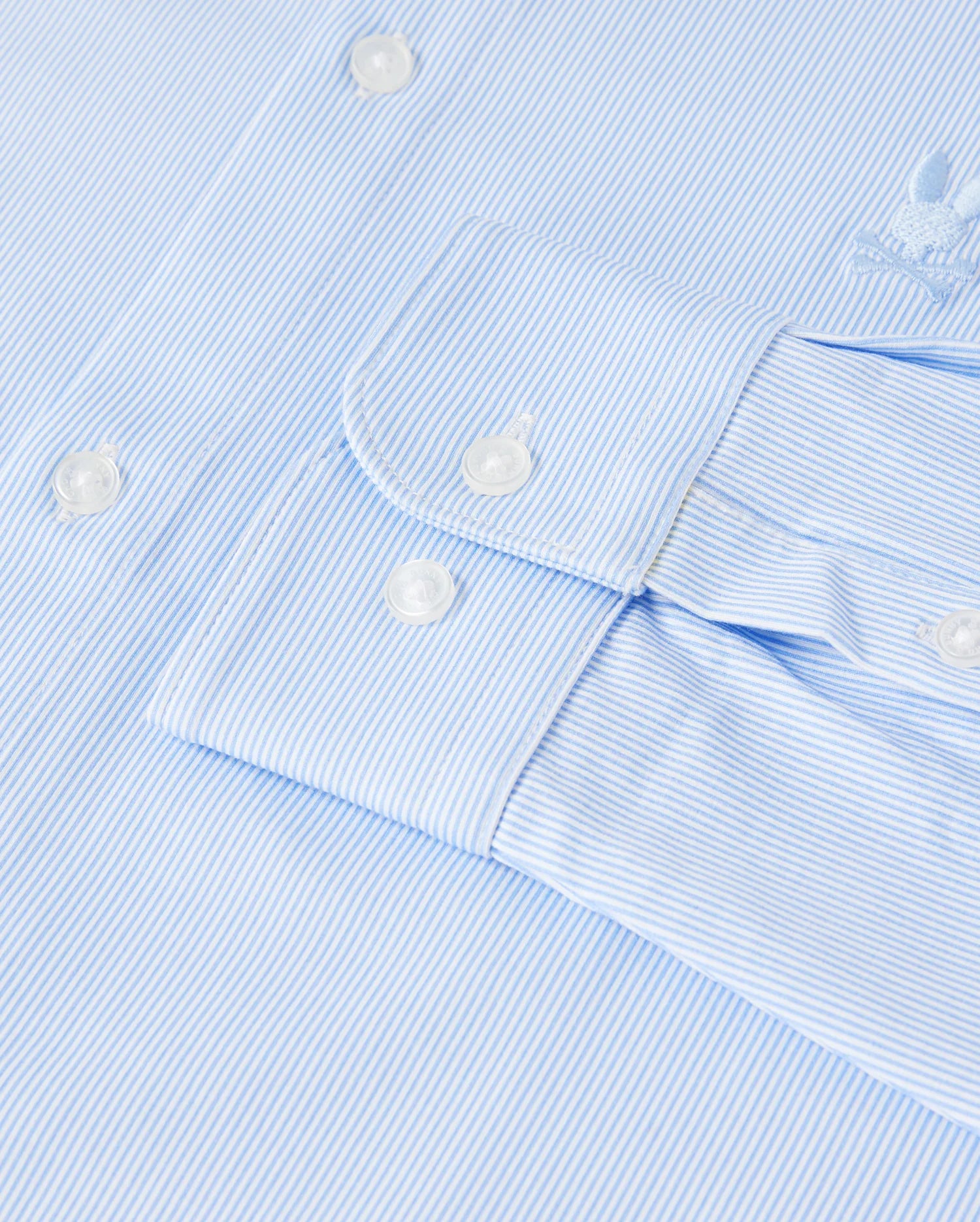 Close-up of a light blue and white pinstripe *Mens Brook Striped Jersey Shirt - B6C750D200* by Psycho Bunny, featuring white buttons on the cuff. The texture of the Pima cotton fabric is clearly visible, with an embroidered logo located near the shoulder. This comfortable shirt is neatly pressed and clean.