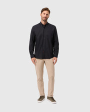 A man stands against a plain background, wearing a Psycho Bunny MENS JARED COTTON MODAL SHIRT in black, beige pants, and green sneakers. His right hand is in his pocket while he maintains a relaxed expression.