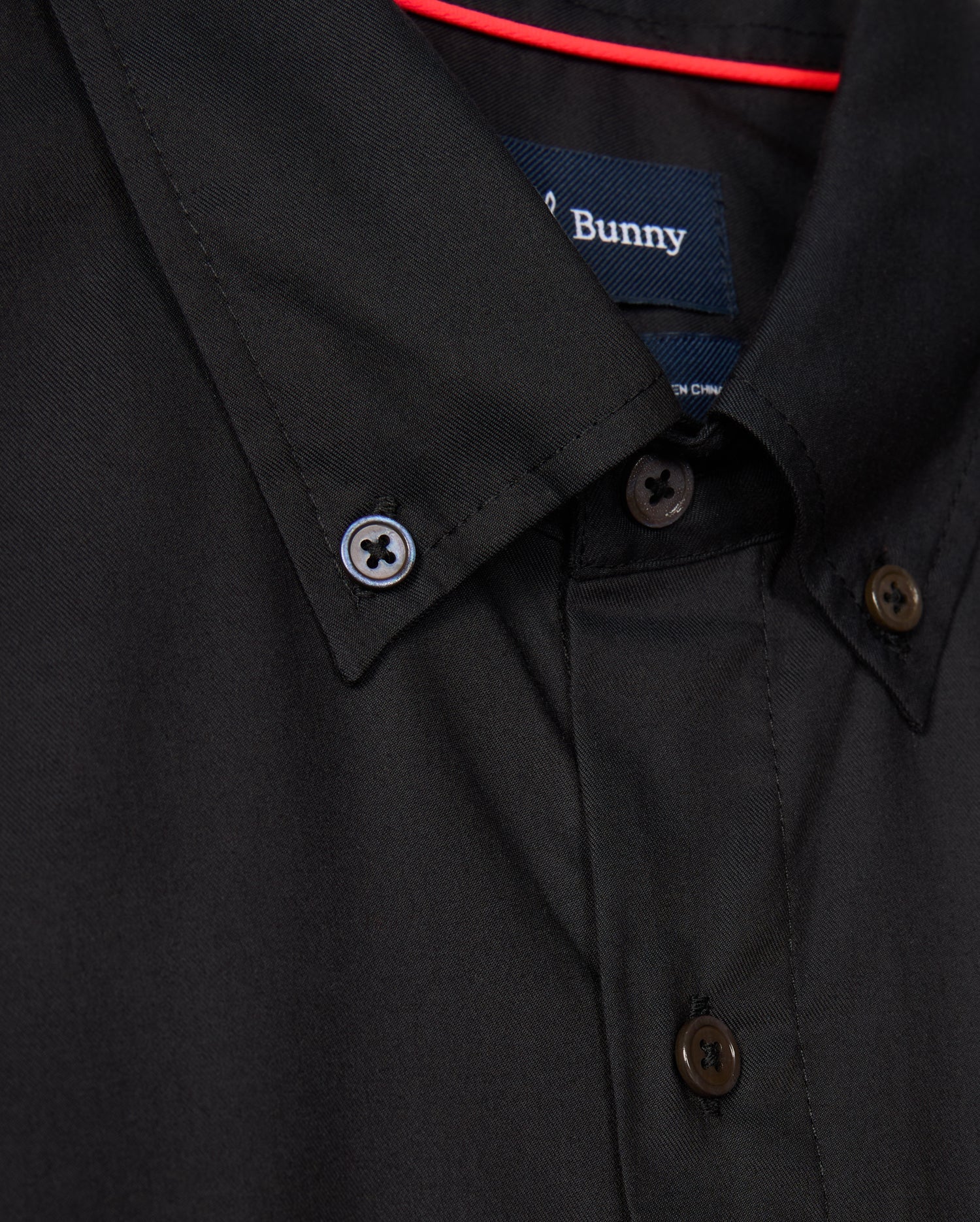 Close-up of the MENS JARED COTTON MODAL SHIRT - B6C774D200 by Psycho Bunny, featuring brown buttons on a premium black fabric. A blue label is sewn inside the collar. A red rod is visible behind the shirt.