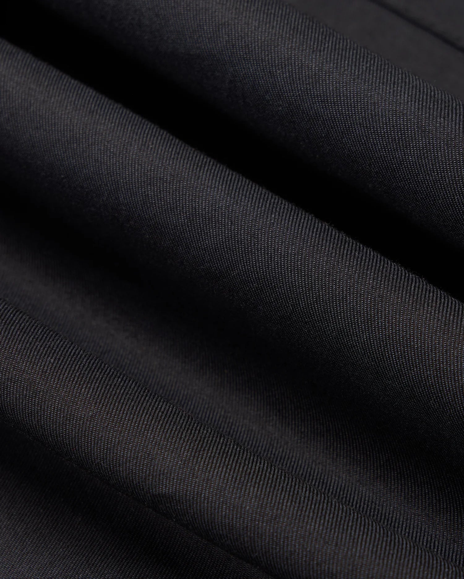 A close-up of Psycho Bunny's MENS JARED COTTON MODAL SHIRT in dark gray or black highlights a premium cotton fabric with subtle diagonal textures, folded slightly to create gentle waves and shadows, ideal for crafting its smooth and soft appearance.