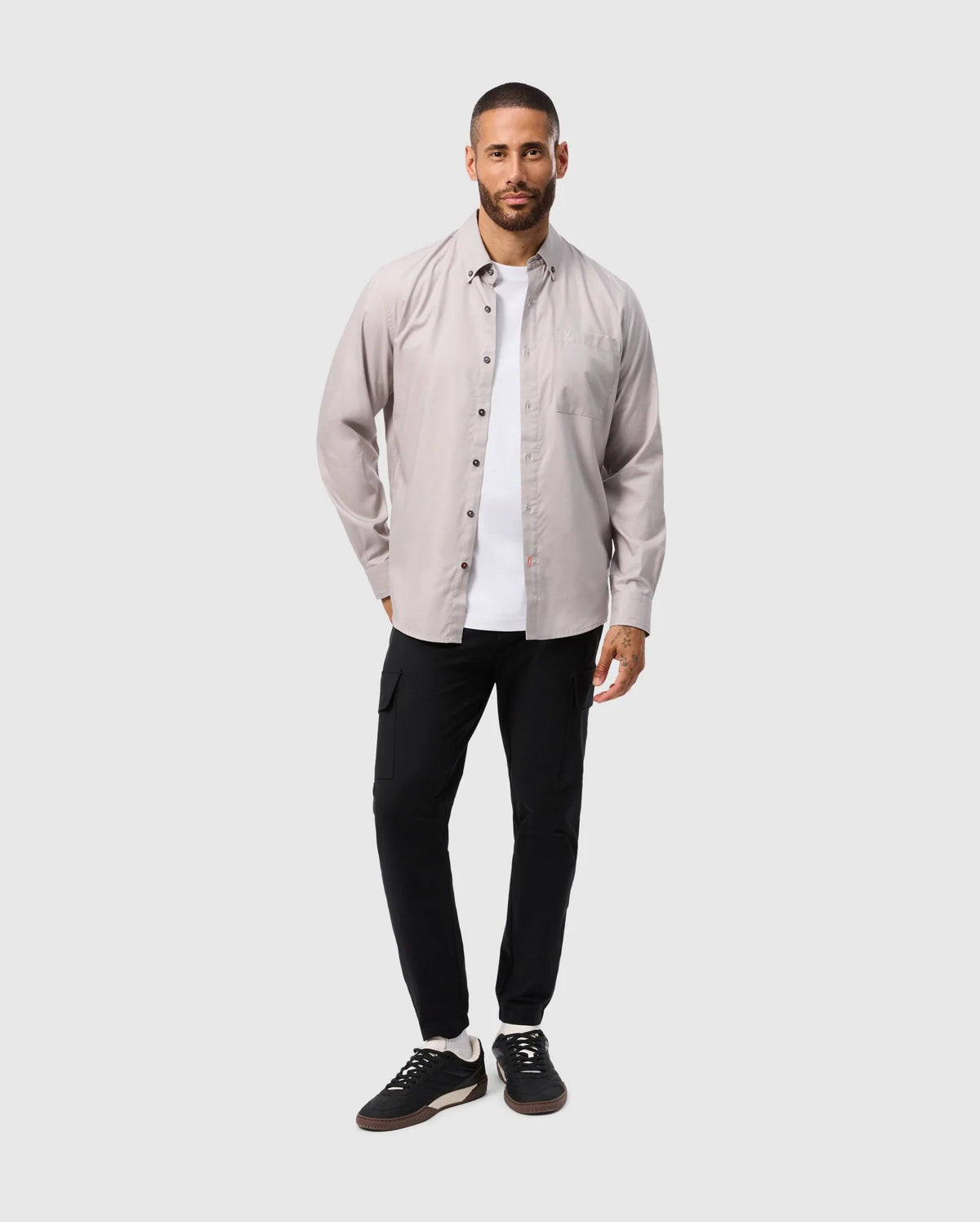 A man with a beard stands against a light gray background, wearing a Psycho Bunny MENS JARED COTTON MODAL SHIRT over a white T-shirt, paired with black pants and sneakers. With his left hand in his pocket, he smiles slightly, exuding casual comfort.