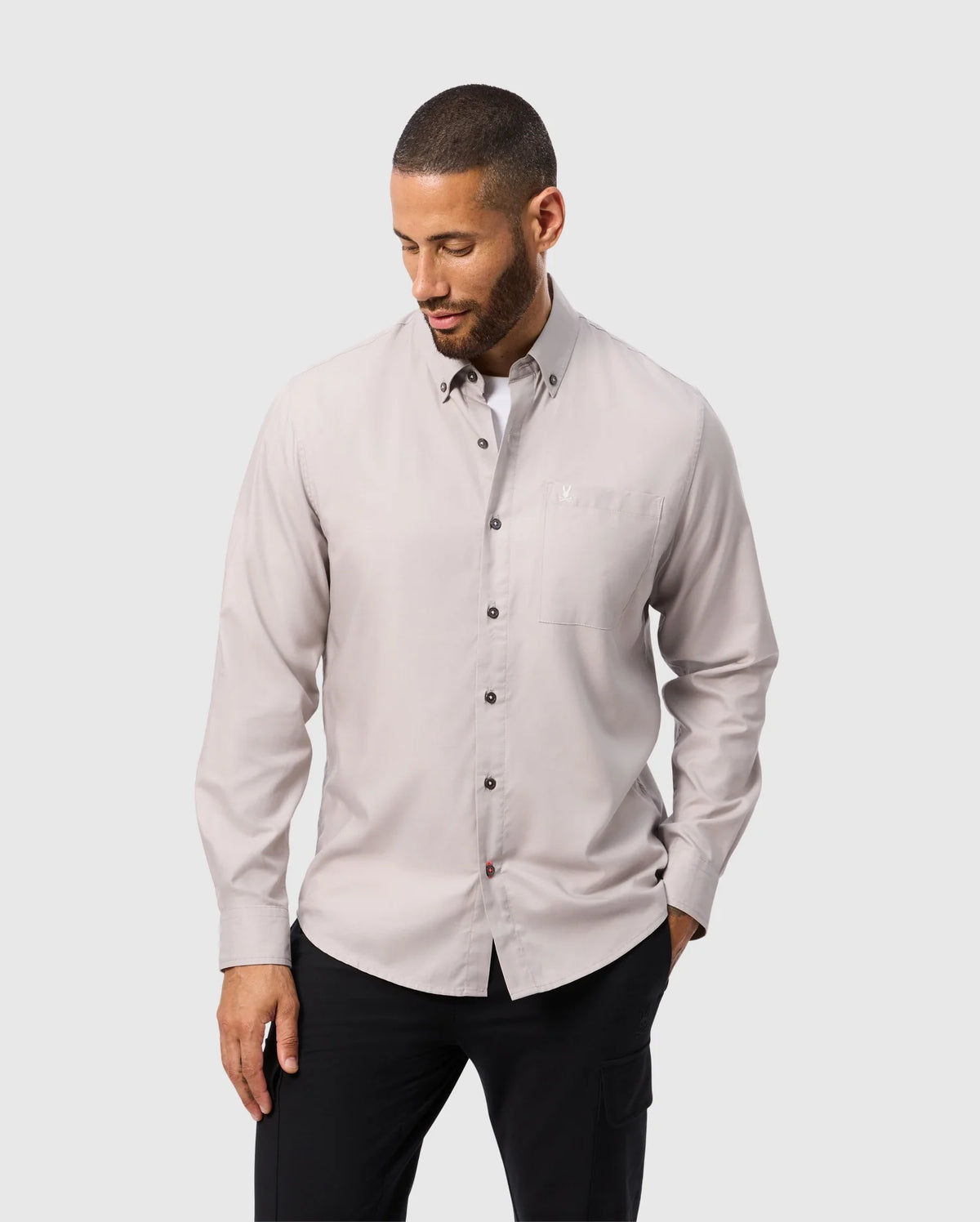 A man in a light gray Psycho Bunny MENS JARED COTTON MODAL SHIRT (B6C774D200) and black pants stands against a plain background. The soft pima cotton modal shirt complements his pose as he looks down, one hand by his side and the other touching his hip.