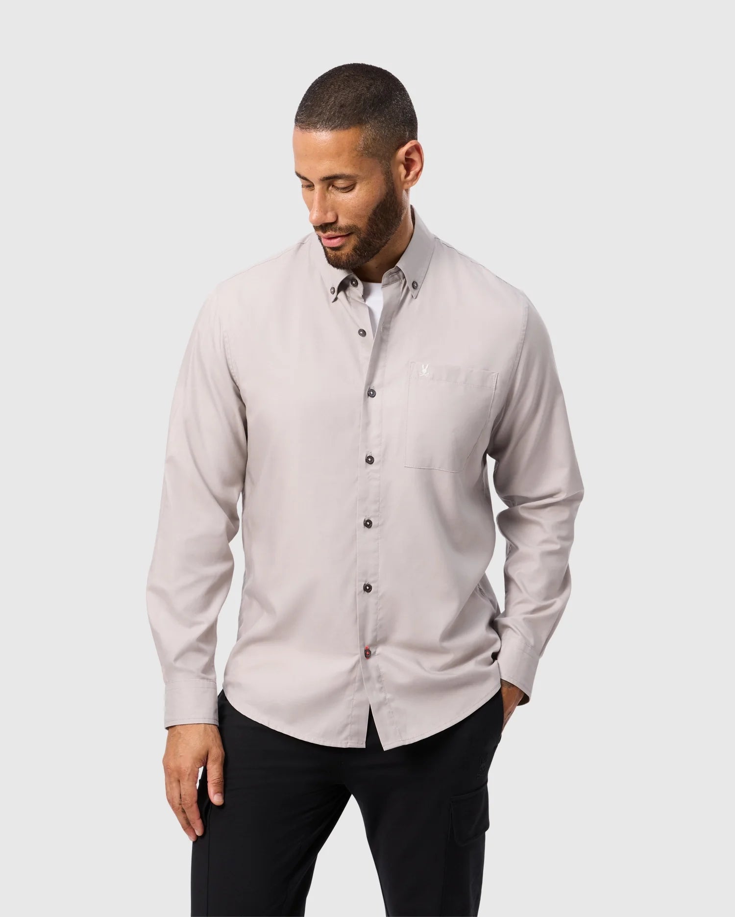 A man in a light gray Psycho Bunny MENS JARED COTTON MODAL SHIRT (B6C774D200) and black pants stands against a plain background. The soft pima cotton modal shirt complements his pose as he looks down, one hand by his side and the other touching his hip.