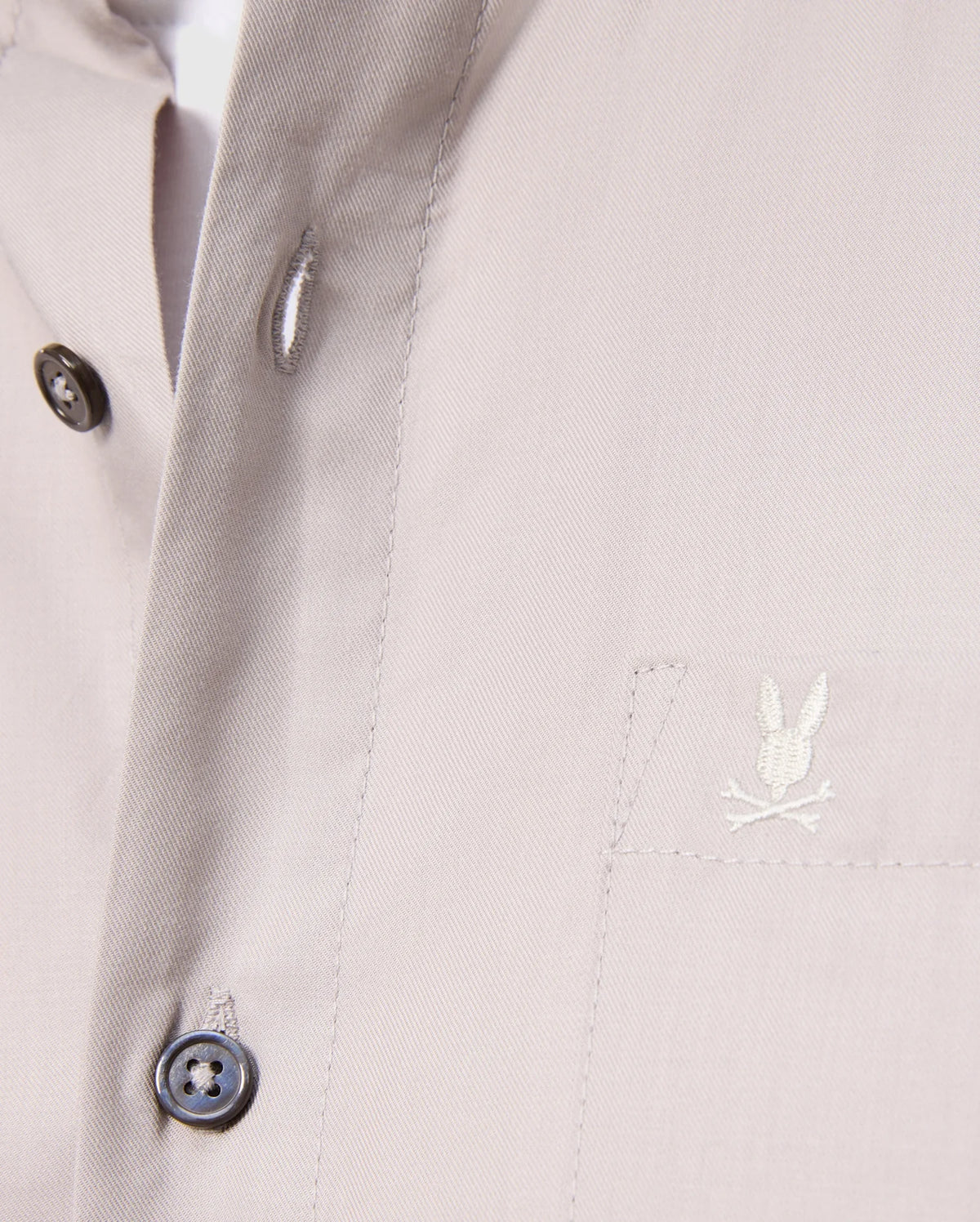 Close-up of a Psycho Bunny Men's Jared Cotton Modal Shirt in beige, with two black placket buttons and an embroidered rabbit logo over crossed bones on the chest pocket. The soft pima cotton modal fabric looks smooth and tailored.