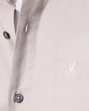 Close-up of a Psycho Bunny Men's Jared Cotton Modal Shirt in beige, with two black placket buttons and an embroidered rabbit logo over crossed bones on the chest pocket. The soft pima cotton modal fabric looks smooth and tailored.