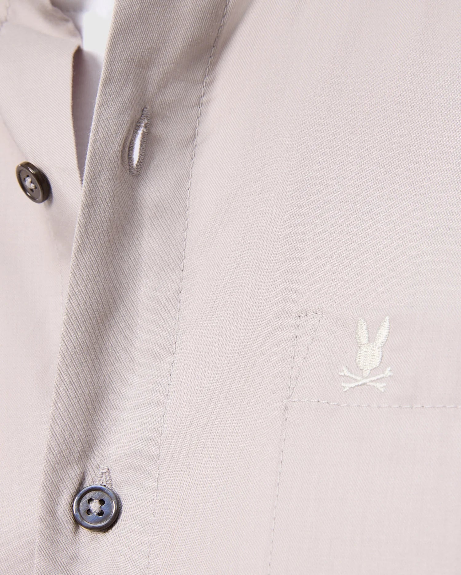 Close-up of a Psycho Bunny Men's Jared Cotton Modal Shirt in beige, with two black placket buttons and an embroidered rabbit logo over crossed bones on the chest pocket. The soft pima cotton modal fabric looks smooth and tailored.