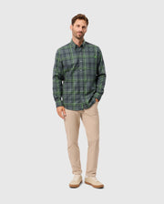 A man with short hair is wearing the Psycho Bunny MENS HAYES PLAID SHIRT in a premium green color, paired with khaki pants. He poses against a plain gray background, looking forward with a neutral expression. His hands rest relaxed at his sides, and he completes the look with white shoes.