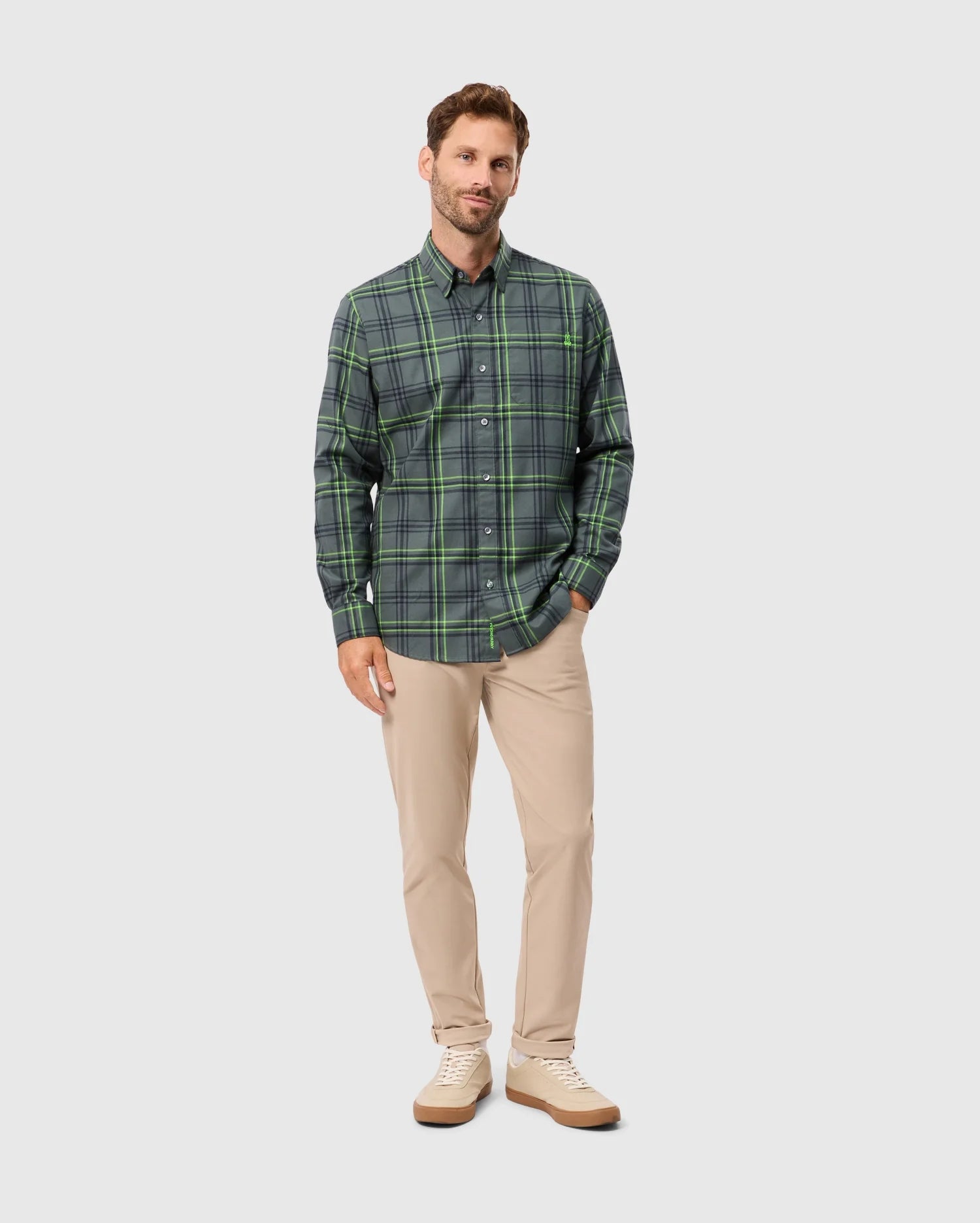 A man with short hair is wearing the Psycho Bunny MENS HAYES PLAID SHIRT in a premium green color, paired with khaki pants. He poses against a plain gray background, looking forward with a neutral expression. His hands rest relaxed at his sides, and he completes the look with white shoes.