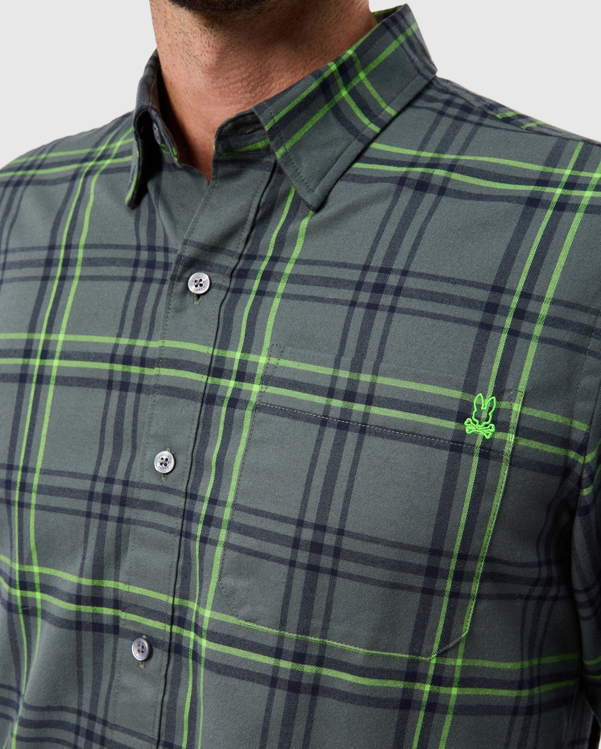 A person is wearing the Psycho Bunny MENS HAYES PLAID SHIRT - B6C842D200, a men's plaid shirt in green and gray with a small green embroidered logo on the chest pocket. This premium button-down shirt features dark buttons and a button-down collar, set against a plain light gray background.