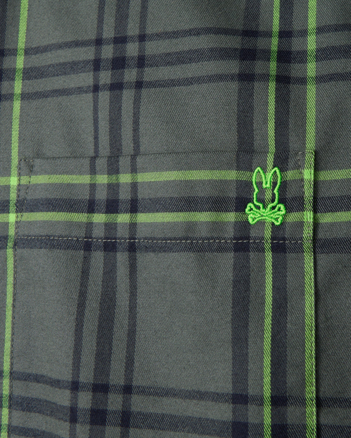 Close-up of the Psycho Bunny Men's Hayes Plaid Shirt - B6C842D200, crafted from premium Pima cotton. The green fabric, featuring intersecting black lines, showcases a small embroidered logo resembling a cartoonish face wearing glasses. The texture and stitching detail of this button-down shirt are clearly visible.