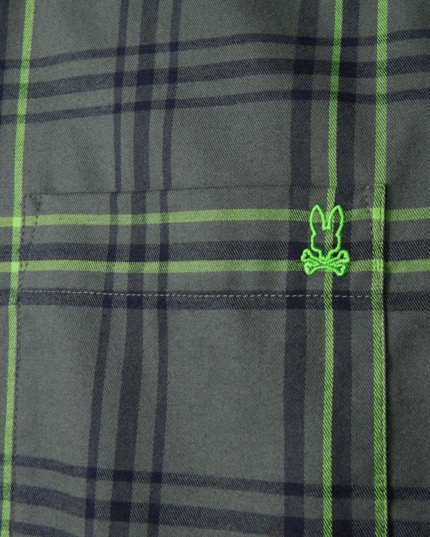Close-up of the Psycho Bunny Men's Hayes Plaid Shirt - B6C842D200, crafted from premium Pima cotton. The green fabric, featuring intersecting black lines, showcases a small embroidered logo resembling a cartoonish face wearing glasses. The texture and stitching detail of this button-down shirt are clearly visible.