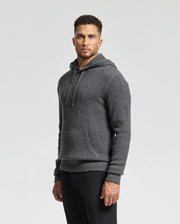 A man in a Psycho Bunny MENS LEVAN WAFFLE HOODED SWEATER - B6E246E200, featuring an embroidered bunny, stands against a plain background. The gray wool-cashmere-blend hoodie has a front pocket and drawstrings. He wears black pants and has short curly hair with a beard, hands at his sides.