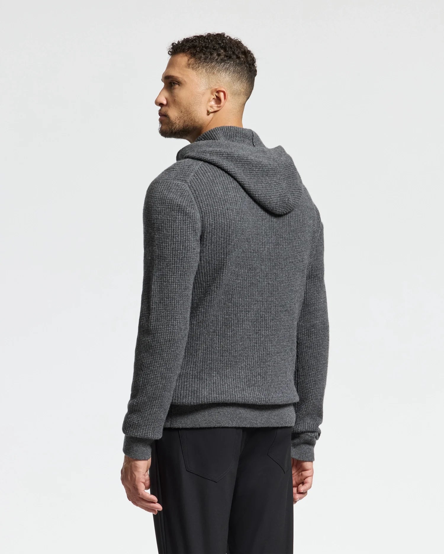 A person with short curly hair and a beard is facing left, wearing a grey Psycho Bunny MENS LEVAN WAFFLE HOODED SWEATER with an embroidered chest bunny and black pants against a plain light background.