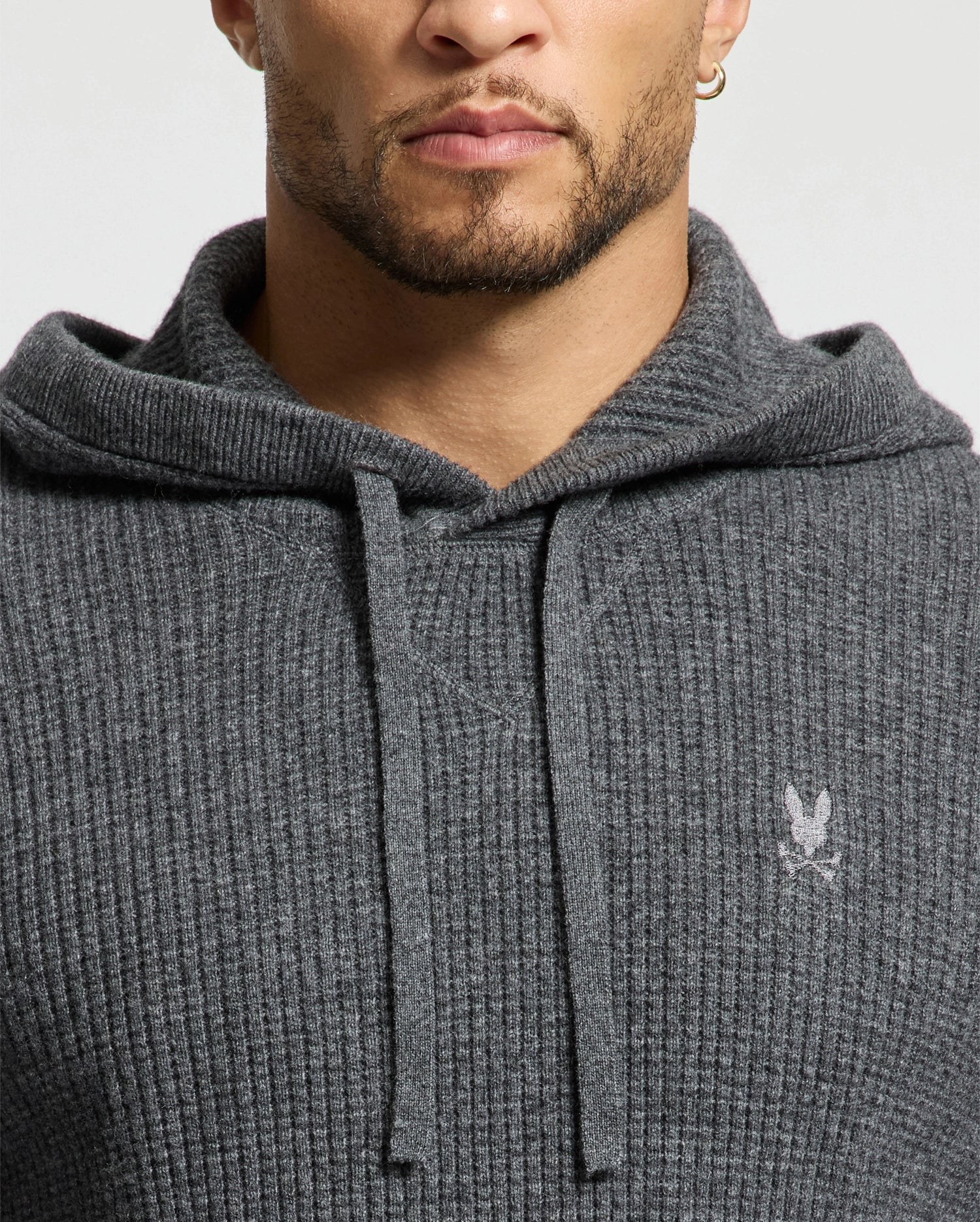 A man wearing a Psycho Bunny gray wool-cashmere-blend hooded MENS LEVAN WAFFLE SWEATER with an embroidered chest bunny, featuring drawstrings. He completes his look with a hoop earring.