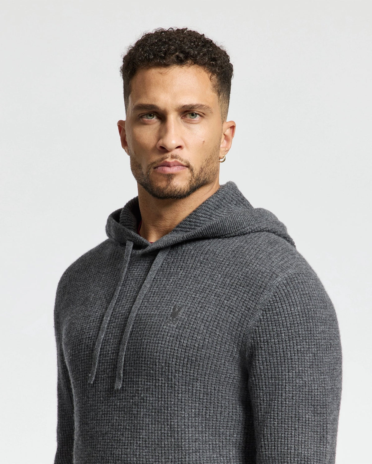 A man with short curly hair wears a Psycho Bunny MENS LEVAN WAFFLE HOODED SWEATER (B6E246E200). He stands against a plain light background, his serious expression complemented by an embroidered chest bunny.