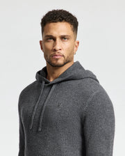 A man with short curly hair wears a Psycho Bunny MENS LEVAN WAFFLE HOODED SWEATER (B6E246E200). He stands against a plain light background, his serious expression complemented by an embroidered chest bunny.