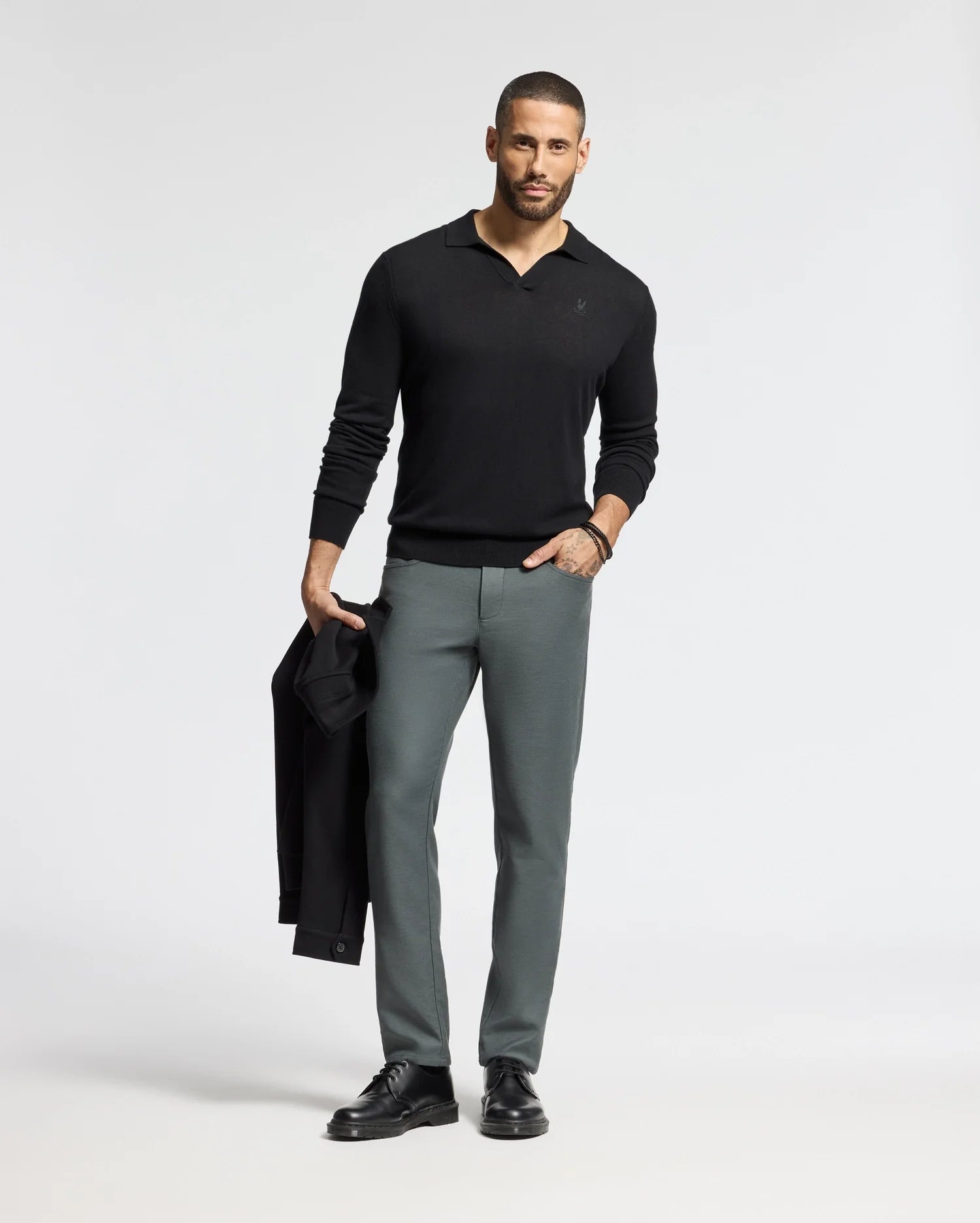 A man with short hair and a beard stands against a plain background, wearing the sleek black MENS JOHNNY COLLAR SWEATER - B6E258E200 by Psycho Bunny and gray pants. He holds a black jacket in one hand, complementing his classic look with black shoes.