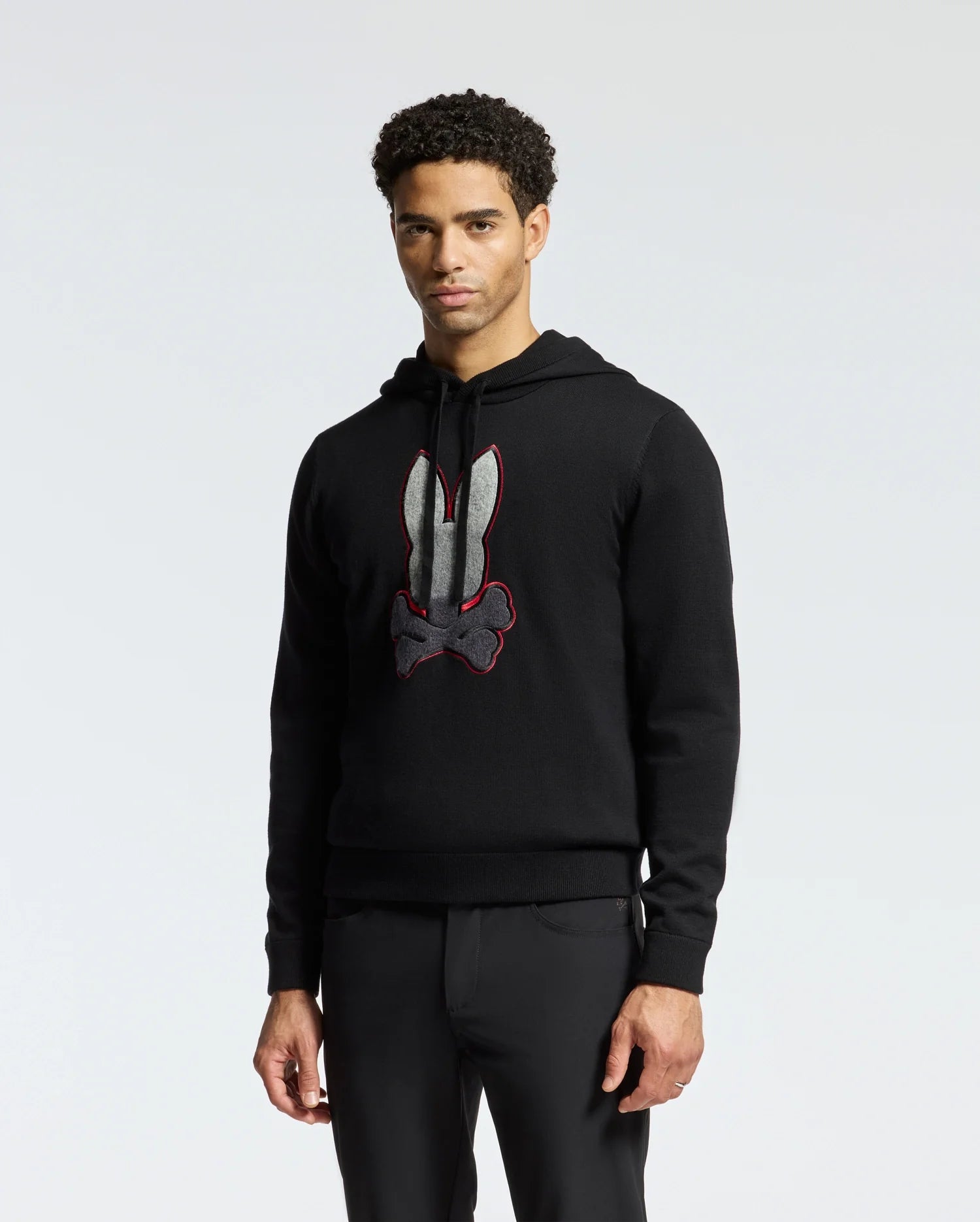 A person is wearing the Psycho Bunny MENS BULDER HOODED SWEATER - B6E261E200, crafted from supple Supima cotton and featuring a large white and red stylized rabbit design on the front. This hoodie is perfectly paired with black pants against a plain white background.
