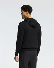 A person wearing the Psycho Bunny MENS BULDER HOODED SWEATER - B6E261E200, made from luxuriously soft Supima cotton, paired with black pants stands against a plain light background, facing slightly away from the camera. The image showcases the person's back and a side profile of their face.