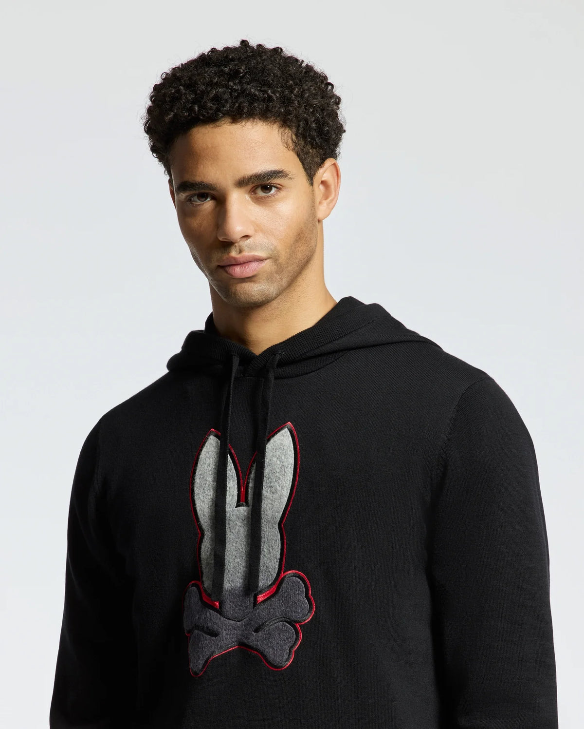 A person wearing the Psycho Bunny MENS BULDER HOODED SWEATER - B6E261E200, designed with a black graphic design of a bunny and crossbones. Made from soft Supima cotton, it elegantly accentuates their curly hair as they look towards the camera against a light gray background.