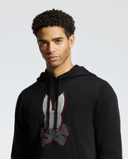 A person wearing the Psycho Bunny MENS BULDER HOODED SWEATER - B6E261E200, designed with a black graphic design of a bunny and crossbones. Made from soft Supima cotton, it elegantly accentuates their curly hair as they look towards the camera against a light gray background.