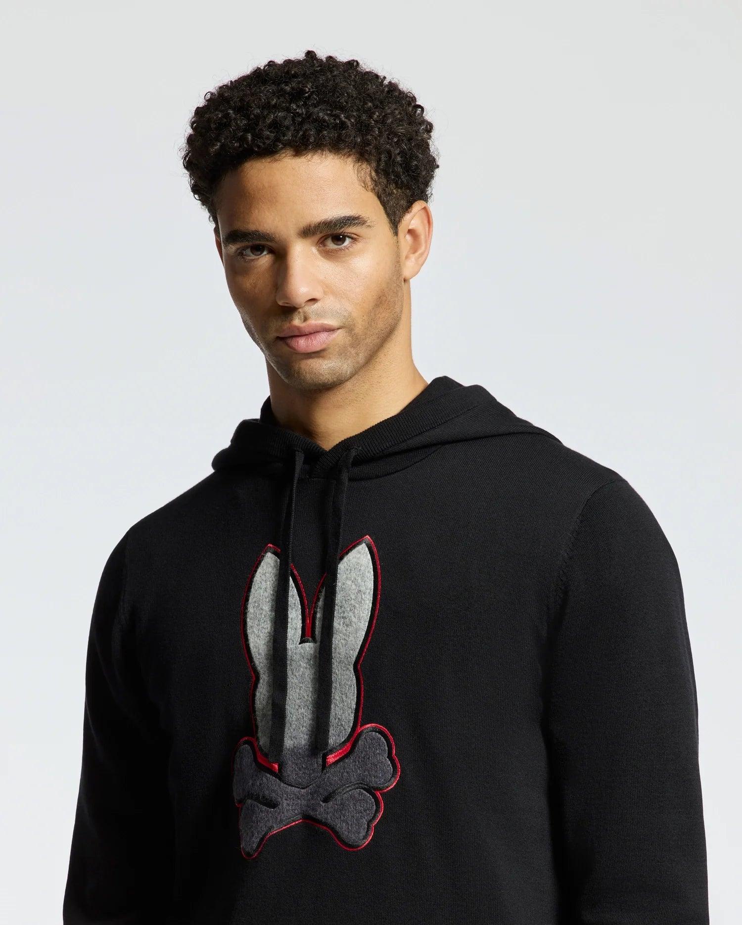 A person wearing the Psycho Bunny MENS BULDER HOODED SWEATER - B6E261E200, designed with a black graphic design of a bunny and crossbones. Made from soft Supima cotton, it elegantly accentuates their curly hair as they look towards the camera against a light gray background.