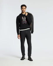 A man stands against a plain background, wearing the MENS BULDER HOODED SWEATER by Psycho Bunny, black pants, and black boots. He holds a dark jacket over his shoulder. His expression is neutral as he faces forward.