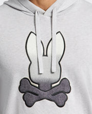 A person wearing a light gray Psycho Bunny MENS BULDER HOODED SWEATER - B6E261E200, featuring a rabbit head above two crossed bones, similar to a skull and crossbones motif.