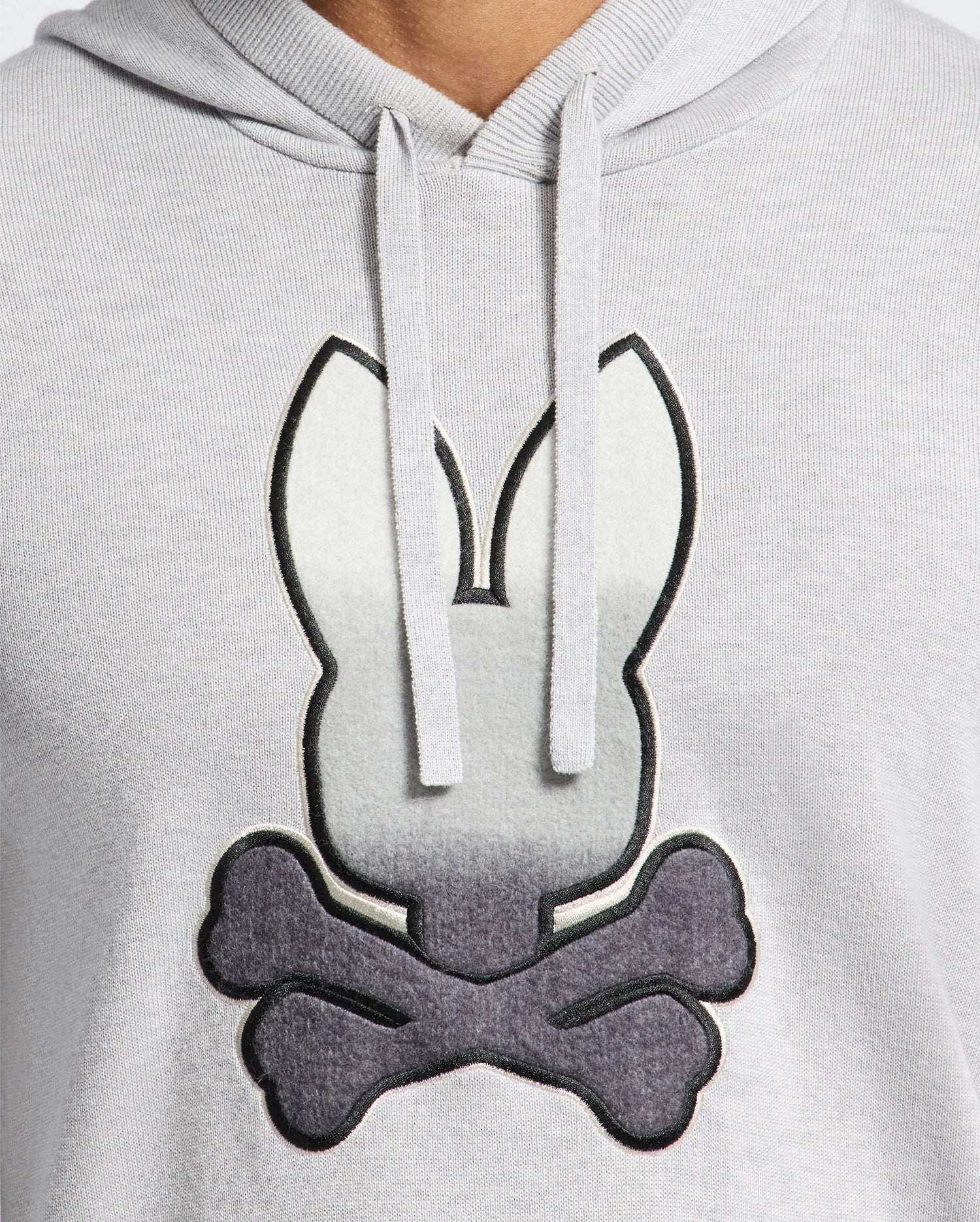 A person wearing a light gray Psycho Bunny MENS BULDER HOODED SWEATER - B6E261E200, featuring a rabbit head above two crossed bones, similar to a skull and crossbones motif.
