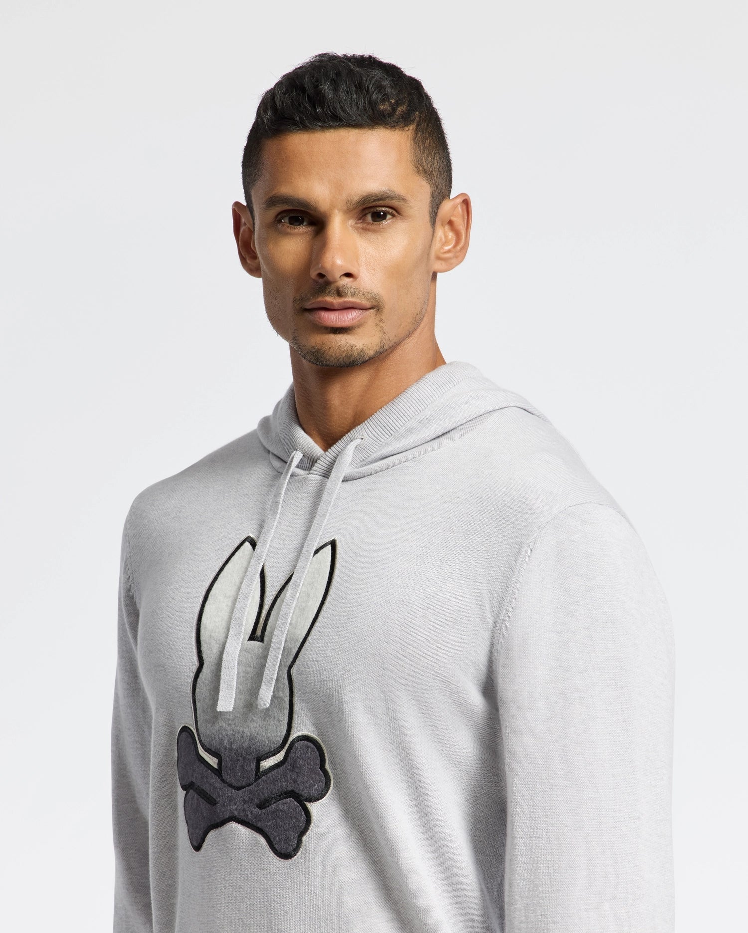 A person with short dark hair poses against a white background, wearing a light gray Psycho Bunny MENS BULDER HOODED SWEATER (B6E261E200), featuring a black and white bunny head and crossbones graphic on the chest.