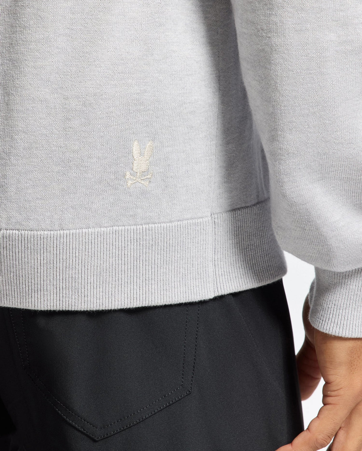 A person wears a Psycho Bunny MENS BULDER HOODED SWEATER - B6E261E200 in light gray, featuring a small embroidered logo on the hem. Paired with dark pants, the image focuses on clothing details with the person partially cropped out.
