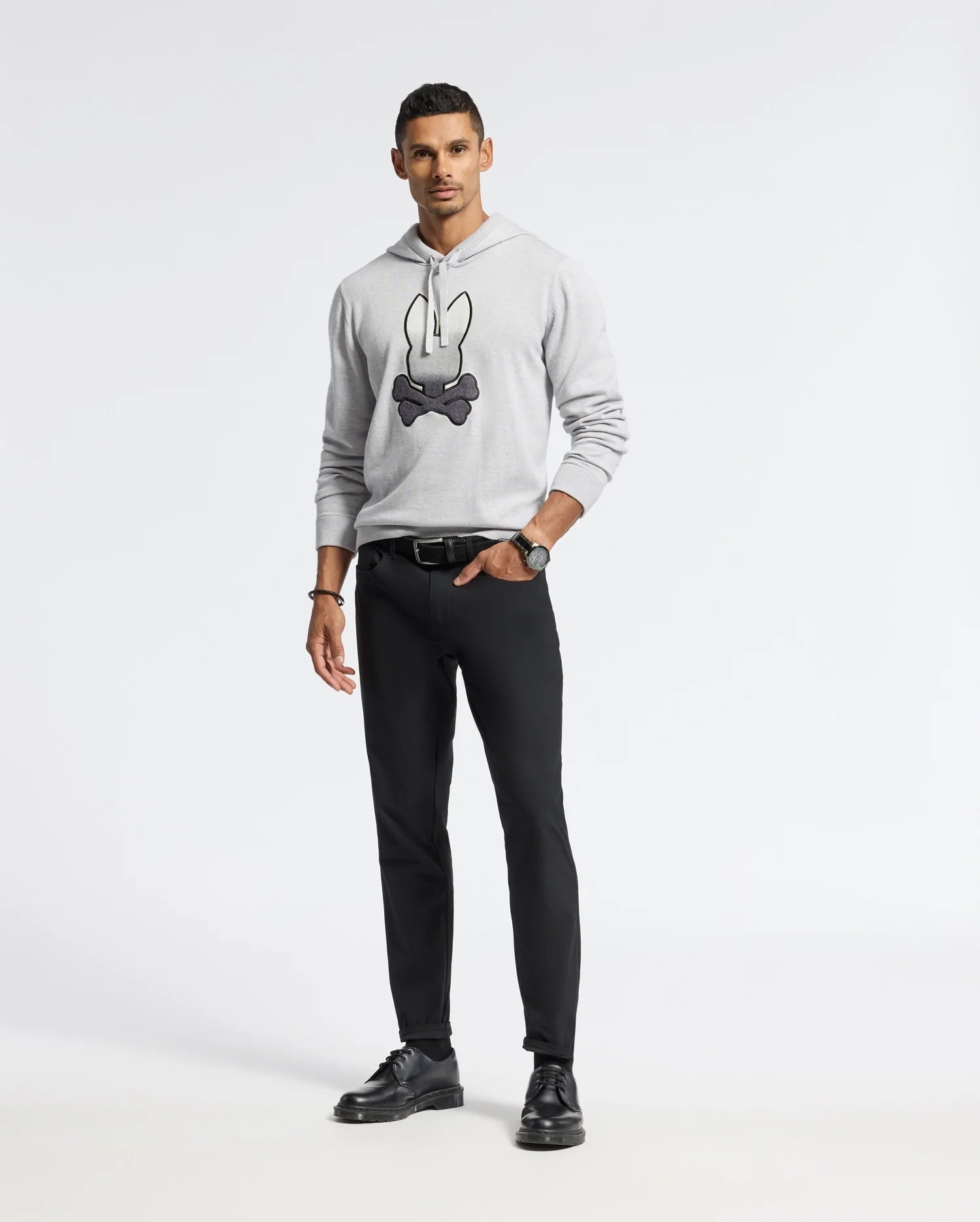 A man wears a Psycho Bunny MENS BULDER HOODED SWEATER in light gray with a cartoon design, paired with black pants. He complements the outfit with black shoes and a watch, posing neutrally against a white background.