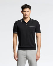 A person stands wearing the Psycho Bunny MENS LONGFORD FINE KNIT POLO - B6E500F200, a black shirt with white trim and an embroidered chest pocket, paired with light gray pants. The fine-knit cotton polo complements the plain white background seamlessly.