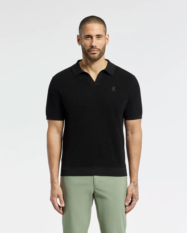 A bearded man confidently wears a breathable Psycho Bunny MENS LEETON JOHNNY COLLAR KNIT POLO (B6E562F200) in black, paired with light green pants. With an embroidered bunny, the 100% cotton shirt fits him perfectly as he gazes directly at the camera, showcasing his left-hand tattoo against a plain backdrop.