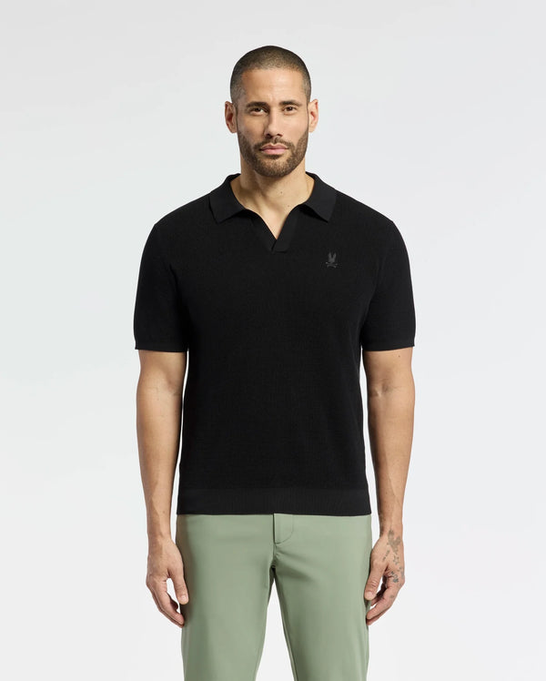 A bearded man confidently wears a breathable Psycho Bunny MENS LEETON JOHNNY COLLAR KNIT POLO (B6E562F200) in black, paired with light green pants. With an embroidered bunny, the 100% cotton shirt fits him perfectly as he gazes directly at the camera, showcasing his left-hand tattoo against a plain backdrop.