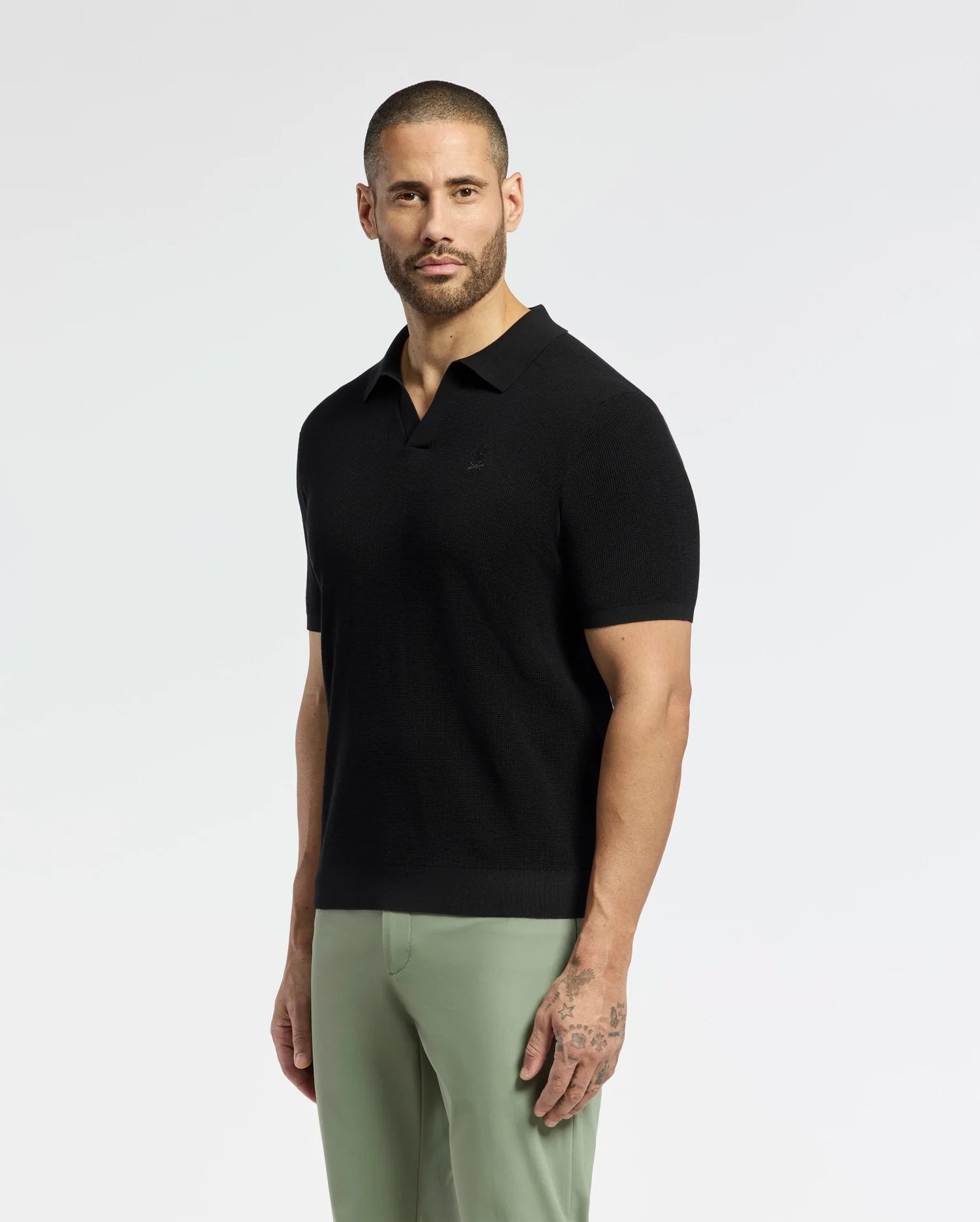 A bearded person stands against a plain backdrop in a black MENS LEETON JOHNNY COLLAR KNIT POLO by Psycho Bunny with light green pants. Their 100% breathable cotton polo features an embroidered Bunny, and tattoos are visible on one arm as they face the camera.